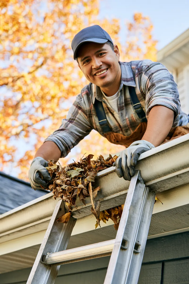 Man on a ladder clearing gutters in autumn, seasonal maintenance behind a $2,500 loan